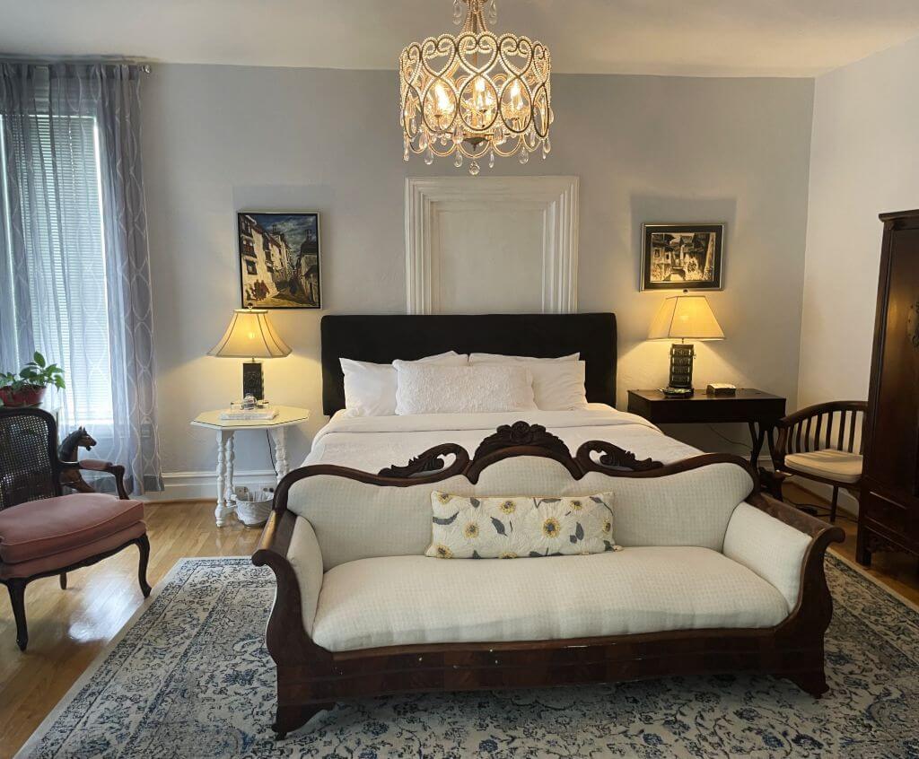 King-size bed with white cozy bedding, wood headboard and a Victorian sitting bench at foot of bed